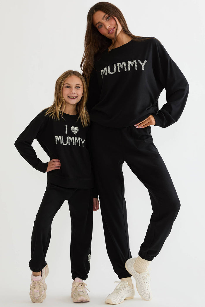 A woman and a girl modeling matching black sweatpants and sweatshirts with white text, standing against a plain white studio background.