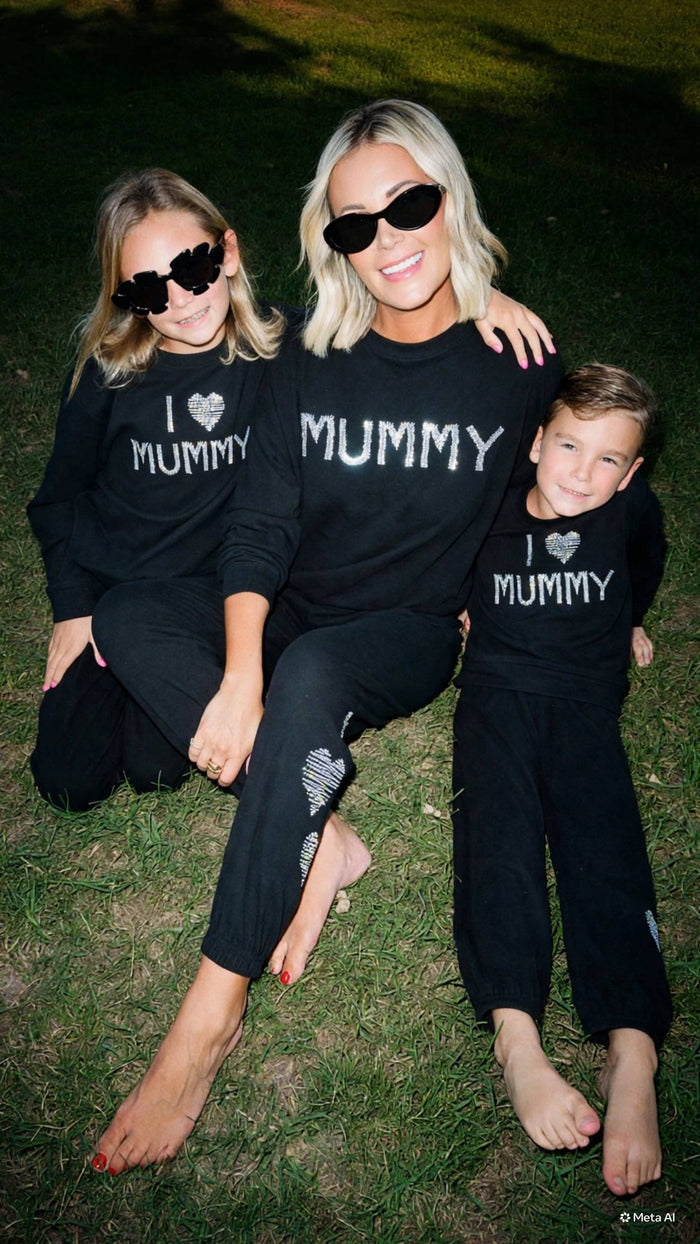 A mother and her two children wearing matching black sweatpants and long-sleeve shirts with 'I [heart] MUMMY' printed in white, posing together on grass at night.