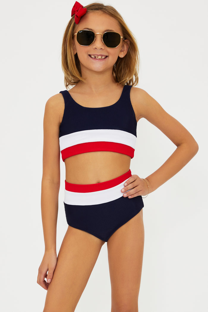 A girl wearing a two-piece swimsuit with an Americana colorblock pattern, consisting of red, white, and blue colors. She is also wearing sunglasses and there is a bow in her hair.