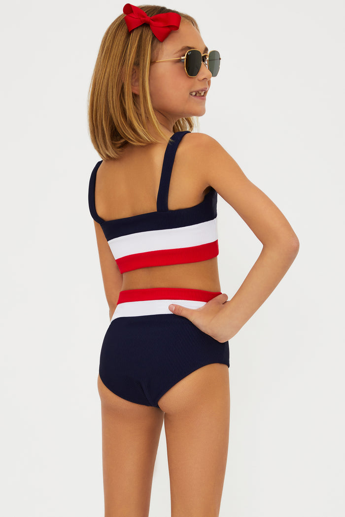 A girl wearing a two-piece swimsuit with an Americana colorblock pattern, consisting of red, white, and blue colors. She is also wearing sunglasses and there is a bow in her hair.