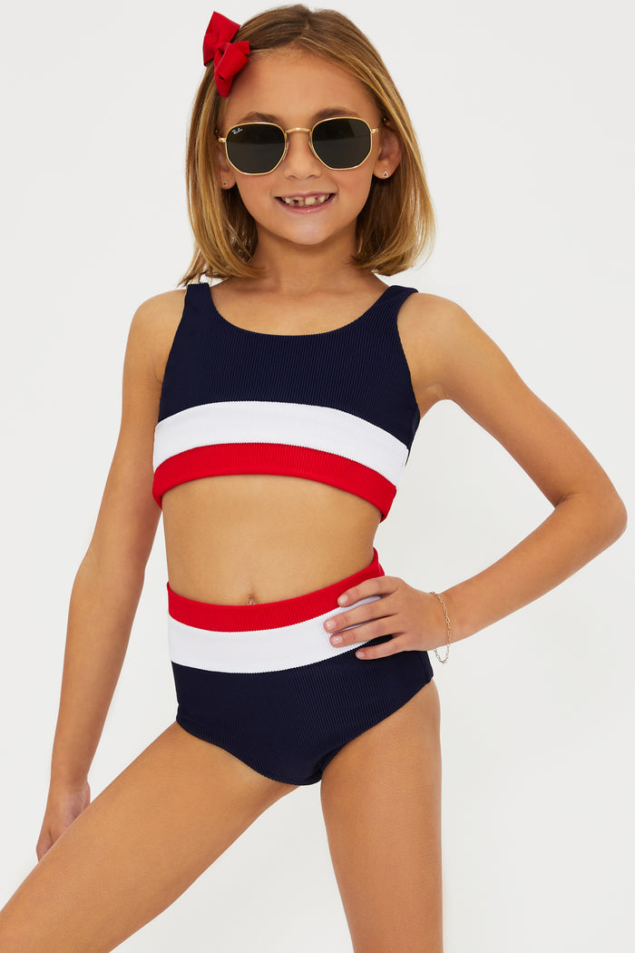 A girl wearing a two-piece swimsuit with an Americana colorblock pattern, consisting of red, white, and blue colors. She is also wearing sunglasses and there is a bow in her hair.