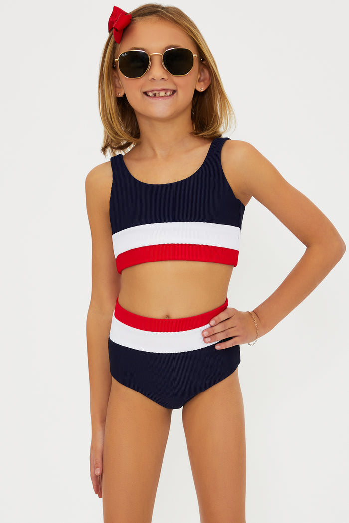 A girl wearing a two-piece swimsuit with an Americana colorblock pattern, consisting of red, white, and blue colors. She is also wearing sunglasses and there is a bow in her hair.