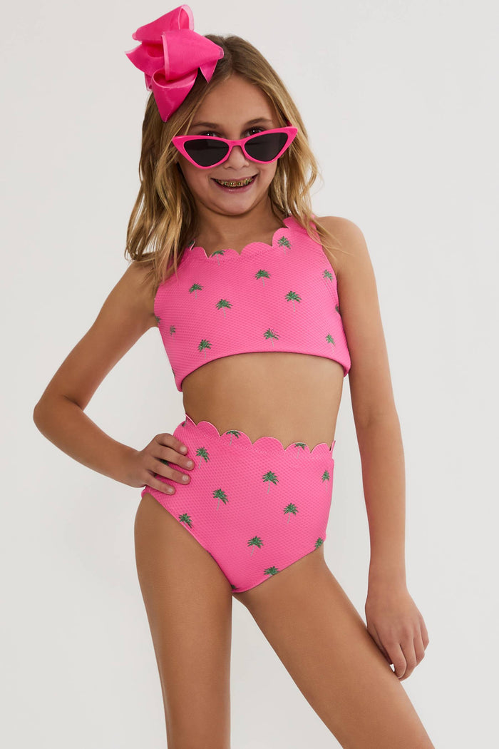 A young girl modeling a bright pink two-piece swimsuit with a small green leaf pattern, matching pink sunglasses, and a large pink bow headband against a light gray studio background.
