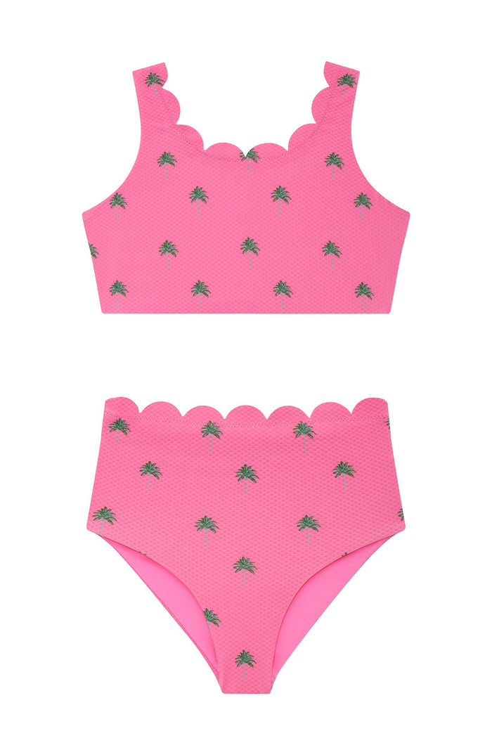 A young girl modeling a bright pink two-piece swimsuit with a small green leaf pattern, matching pink sunglasses, and a large pink bow headband against a light gray studio background.