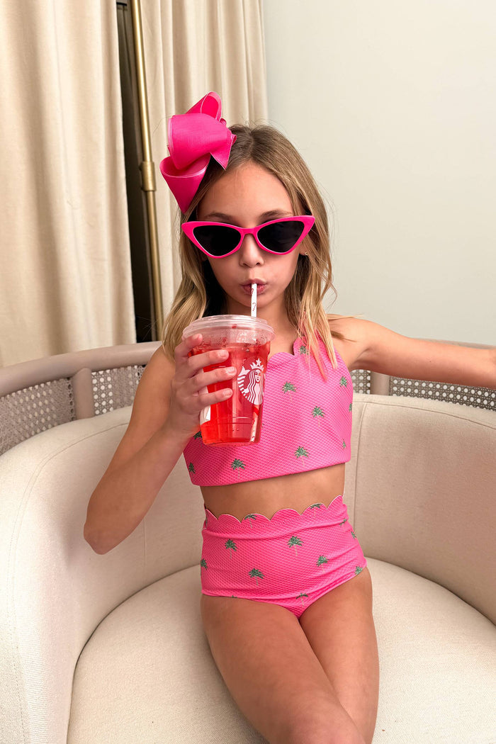 A young girl wearing a bright pink two-piece swimsuit with small green palm tree prints, a large pink bow in her hair, and pink cat-eye sunglasses, sitting on a cream-colored sofa and holding a drink with a Starbucks straw.