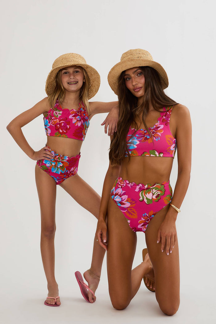 Two women wearing matching pink floral bikini sets and straw hats on a white background
