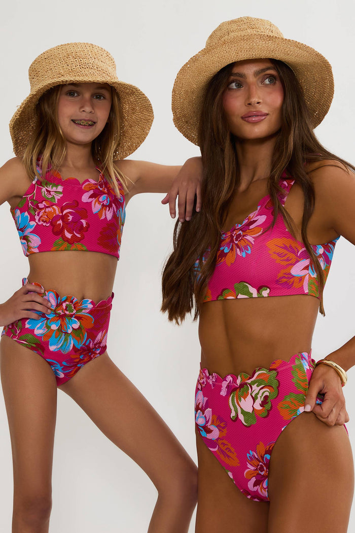 Two women wearing matching pink floral bikini sets with straw hats on a white background