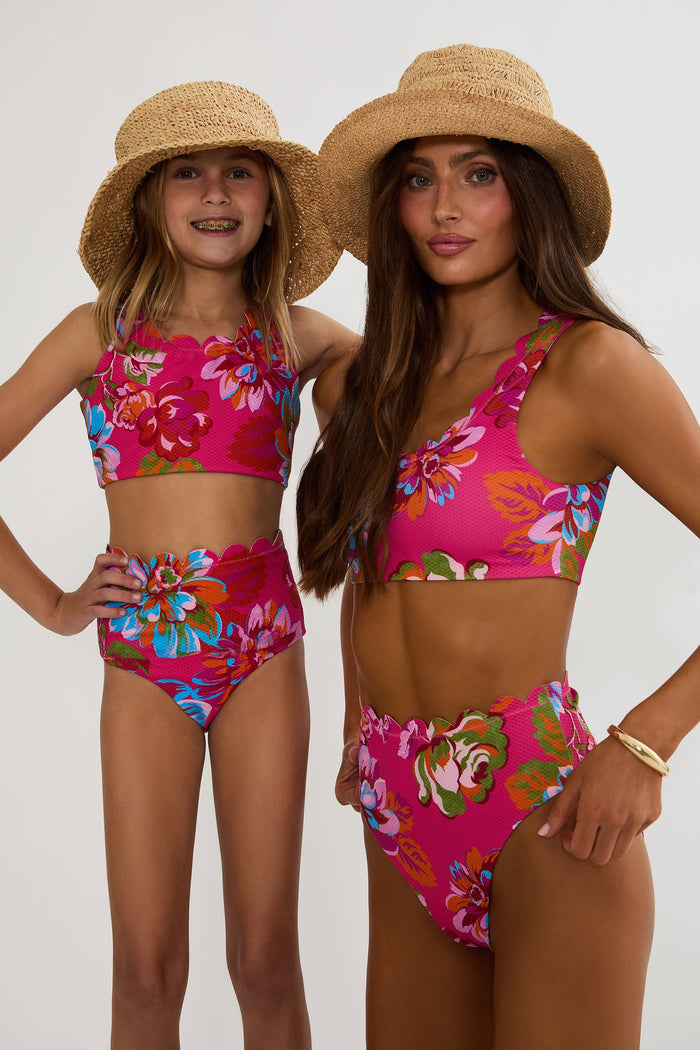 Two women wearing matching pink floral bikini sets with straw hats on a white background
