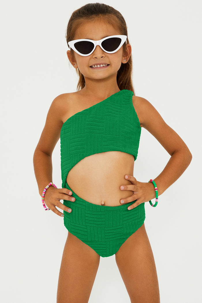 A child model wearing a one-shoulder asymmetrical green swimsuit with a side cut-out detail, paired with sunglasses and colorful bracelets.