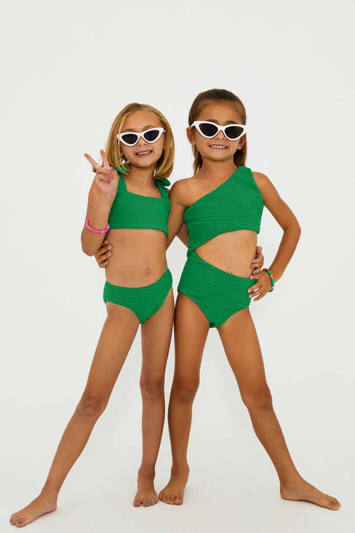 A child model wearing a one-shoulder asymmetrical green swimsuit with a side cut-out detail, paired with sunglasses and colorful bracelets.