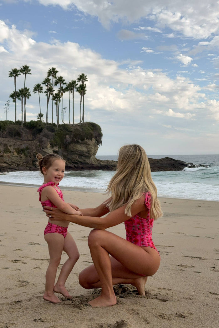 Woman and child in matching pink swimsuits on a beach with palm trees and ocean in the background