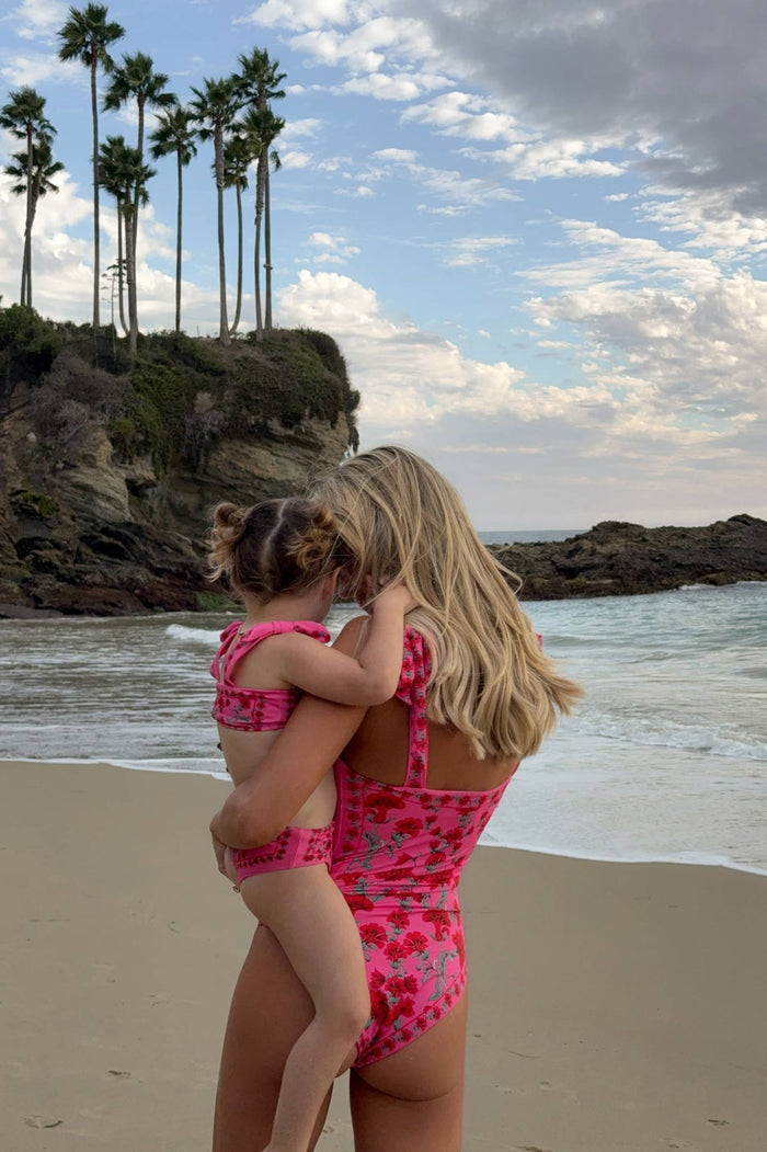 Woman and child in matching pink swimsuits on a beach with palm trees and ocean in the background