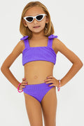 A girl wearing a two-piece swimsuit in ultra violet color with bows on the straps, sunglasses, and standing against a white background.