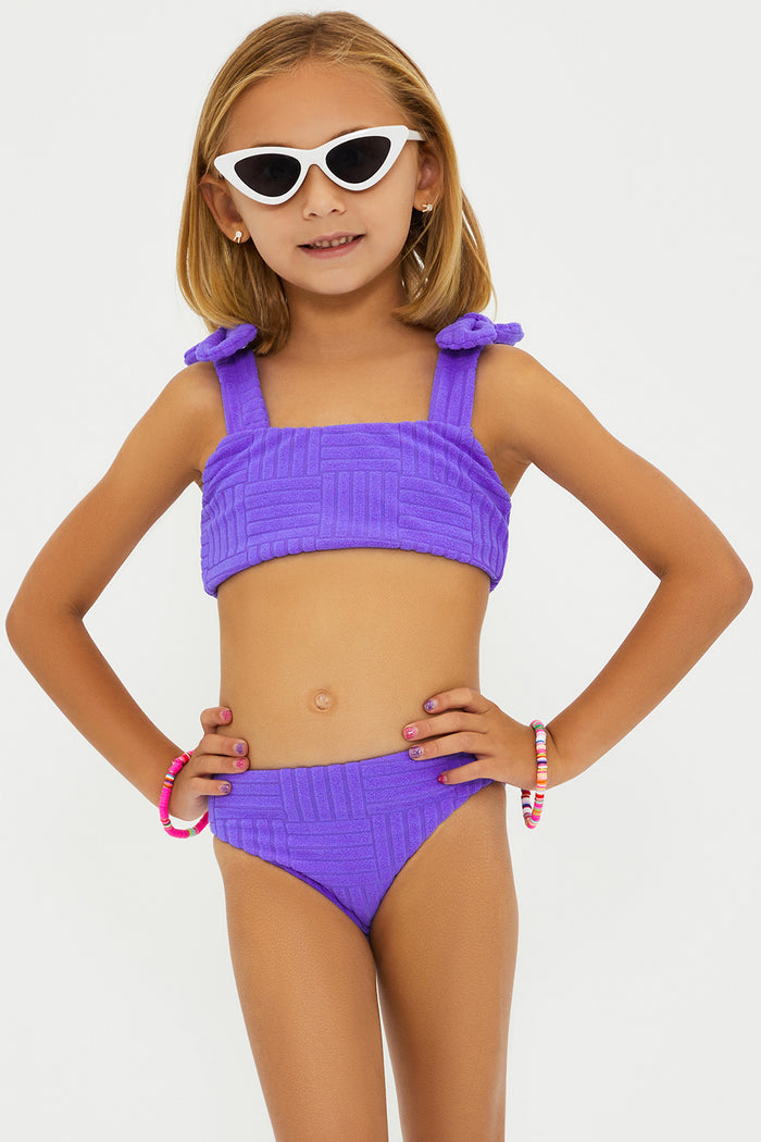 A girl wearing a two-piece swimsuit in ultra violet color with bows on the straps, sunglasses, and standing against a white background.