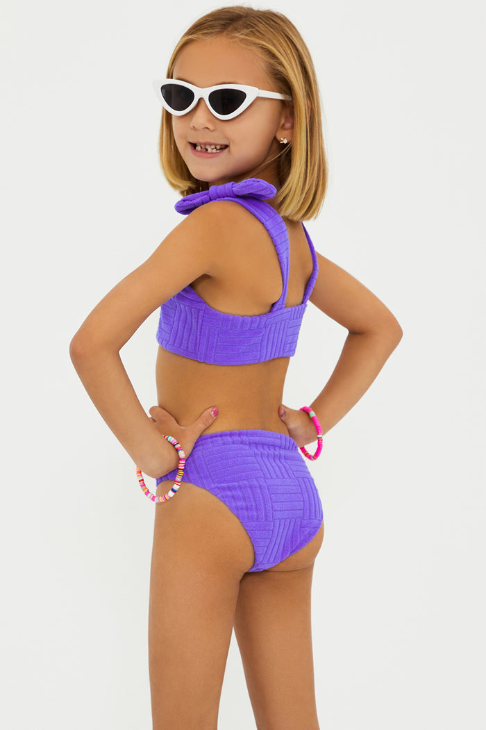 A girl wearing a two-piece swimsuit in ultra violet color with bows on the straps, sunglasses, and standing against a white background.