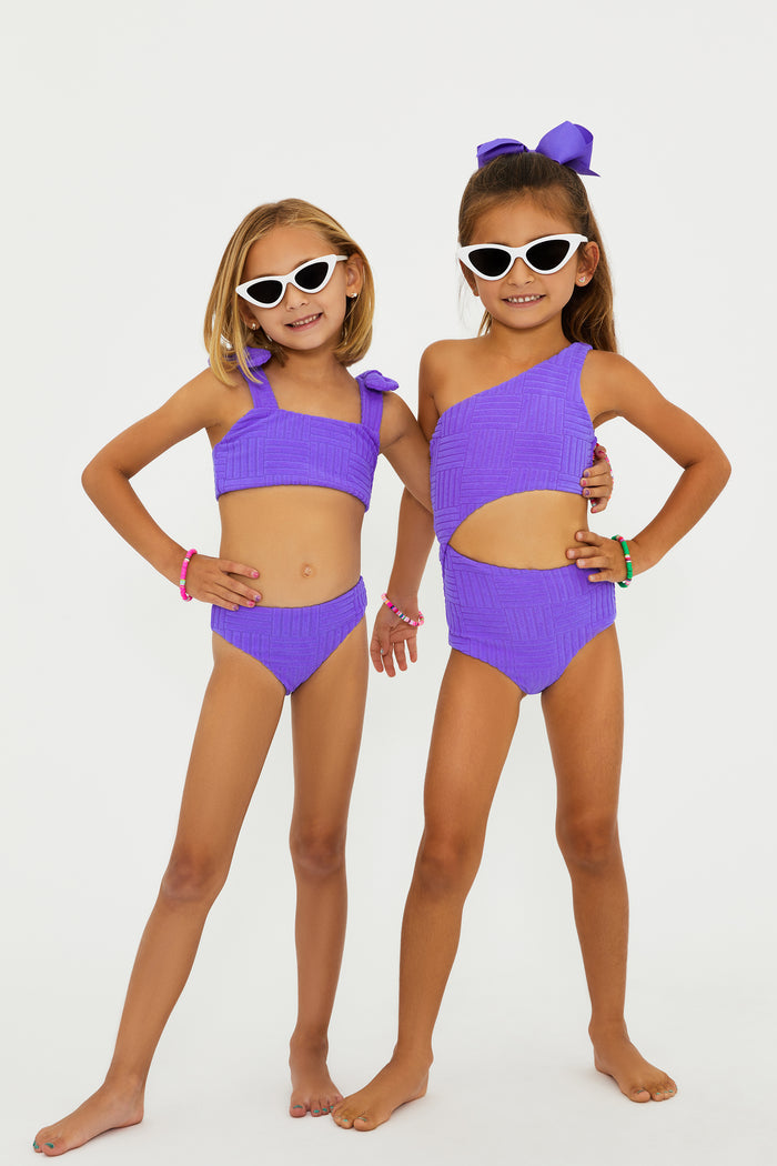 A girl wearing a two-piece swimsuit in ultra violet color with bows on the straps, sunglasses, and standing against a white background.