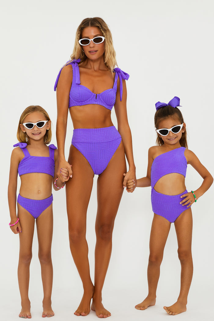 A girl wearing a two-piece swimsuit in ultra violet color with bows on the straps, sunglasses, and standing against a white background.