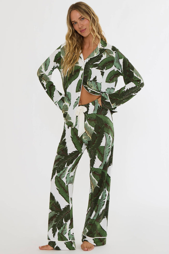 A woman modeling a matching two-piece pajama set with a green tropical leaf print on a white background, featuring a long-sleeve button-up top and wide-leg pants.