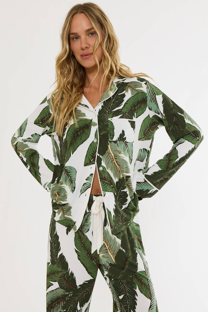 A woman modeling a matching two-piece pajama set with a green tropical leaf print on a white background, featuring a long-sleeve button-up top and coordinating pants.
