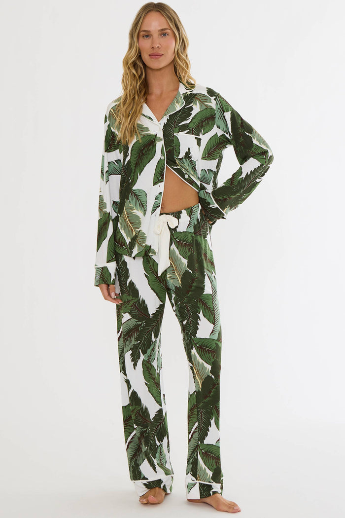 A woman modeling a matching two-piece pajama set with a green tropical leaf print on a white background, featuring a long-sleeve button-up shirt and matching pants.