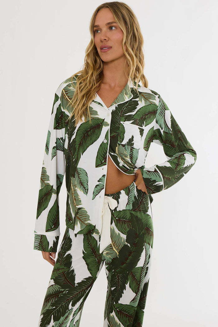 A woman modeling a matching two-piece pajama set with a green tropical banana leaf print on a white background, featuring a long-sleeve button-up top and matching pants.