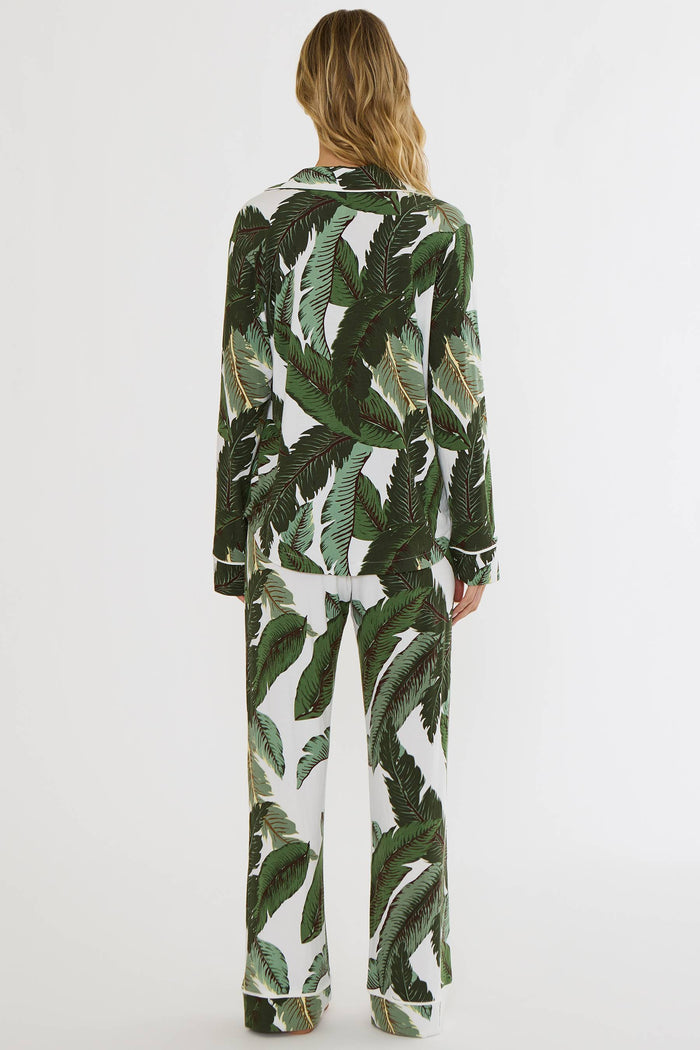 A woman viewed from behind wearing a matching long-sleeve pajama set with a green tropical leaf print on a white background, set against a plain white studio backdrop.