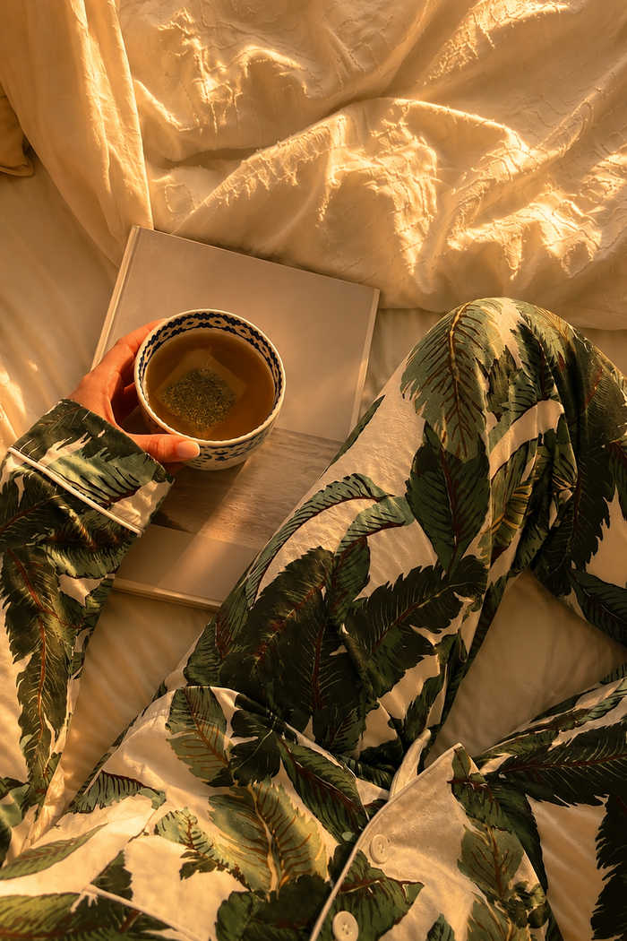 A person wearing green tropical leaf print pajamas relaxes on a bed, holding a cup of tea next to a book in warm golden-hour sunlight.