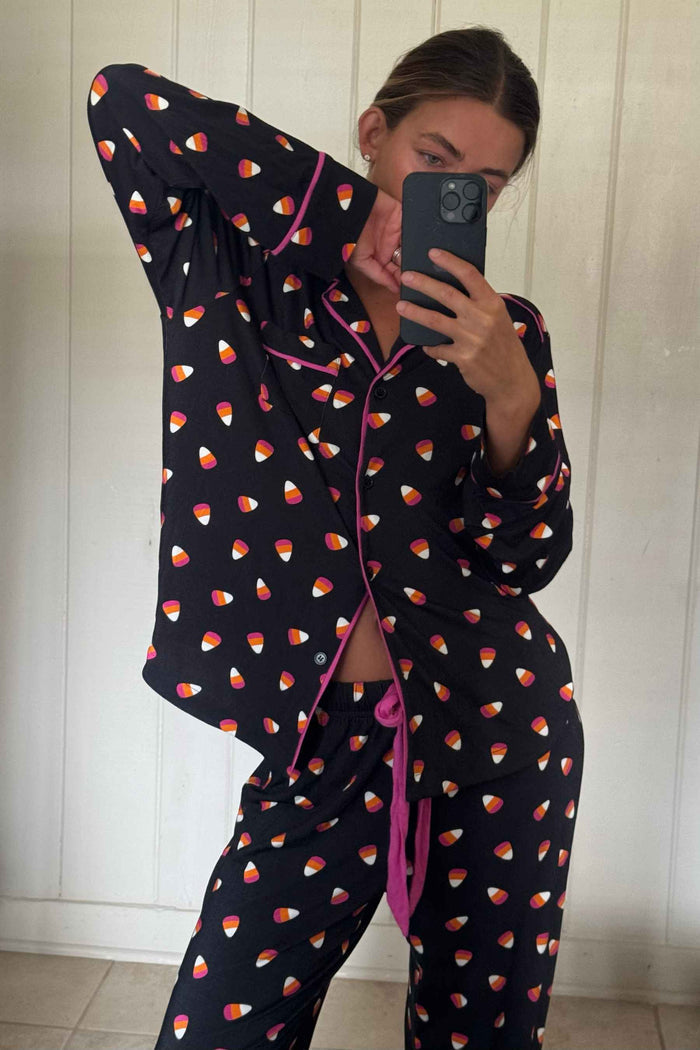 A woman takes a mirror selfie wearing a two-piece pajama set with a black base, colorful candy corn pattern, and bright pink piping and tie details. The background is a white paneled wall.