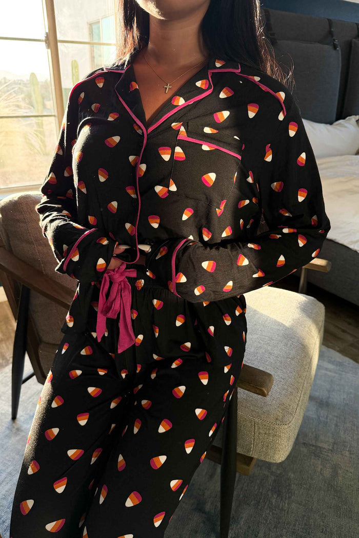 A woman wearing a black long-sleeve pajama set with a candy corn print and pink trim, sitting in a chair in a bedroom setting.