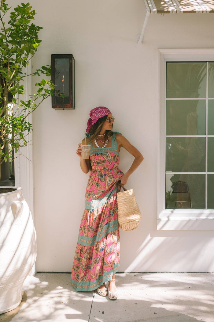 A woman modeling a sleeveless tiered maxi dress with a vibrant pink paisley pattern and turquoise green trim