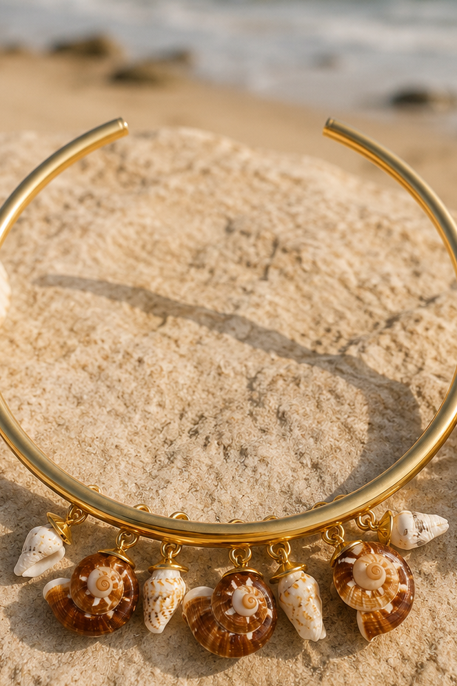 Gold-toned open cuff bracelet with dangling seashell charms, photographed on sand with the ocean blurred in the background.