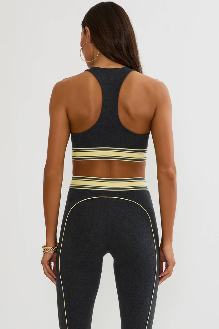 Rear view of a woman wearing a dark charcoal gray athletic set with beige and yellow stripes, consisting of a sports bra and high-waisted leggings, against a light gray studio background.