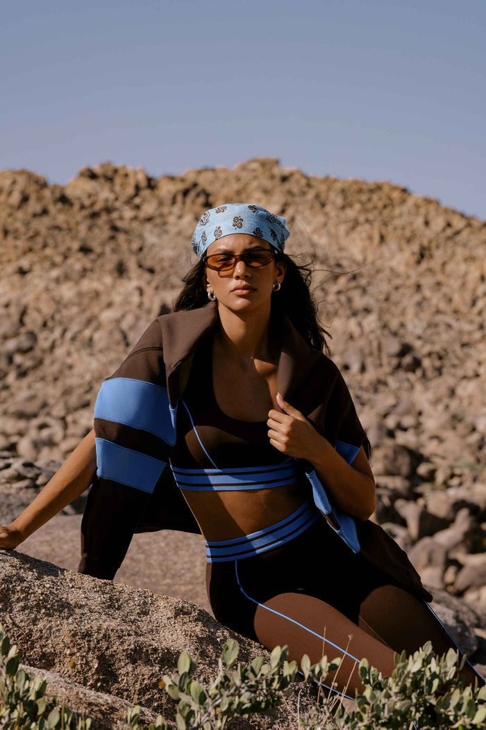 A woman posing outdoors in a rocky desert landscape, wearing a matching brown and blue athletic set with a blue patterned bandana and sunglasses.