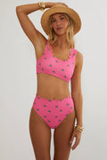 A woman modeling a bright pink two-piece swimsuit with a small green leaf pattern and scalloped edges, styled with a wide-brimmed straw hat and gold bracelets against a light gray studio background.