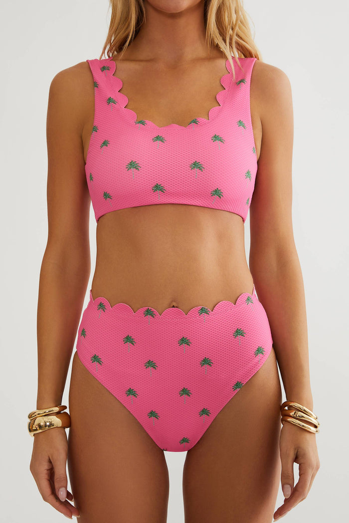 A woman wearing a bright pink two-piece bikini with a small green palm tree pattern and scalloped edges, shown against a light gray studio background.