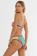 A woman modeling a colorful crocheted bikini set against a plain light gray studio background. The bikini features a bright pink top with green, yellow, and black striped trim, and matching bottoms with pink, green, yellow, and maroon striped details.