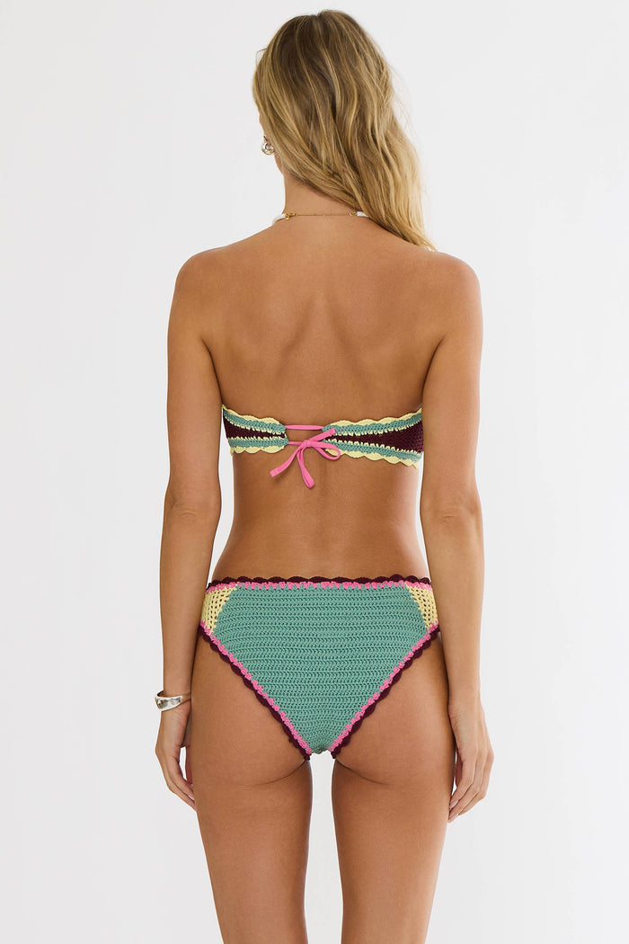 A woman modeling a colorful crocheted bikini set against a plain light gray studio background. The bikini features a bright pink top with green, yellow, and black striped trim, and matching bottoms with pink, green, yellow, and maroon striped details.