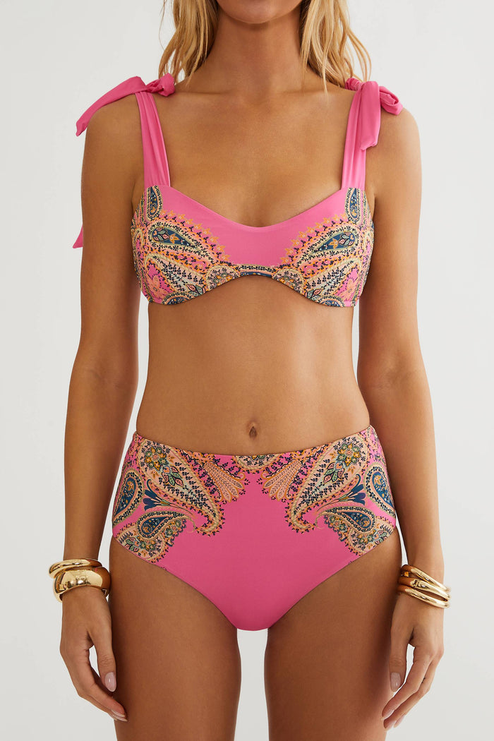 A woman modeling a two-piece bikini with a bright pink base and a multicolor paisley pattern, shown from a side-back angle against a plain light gray studio background.