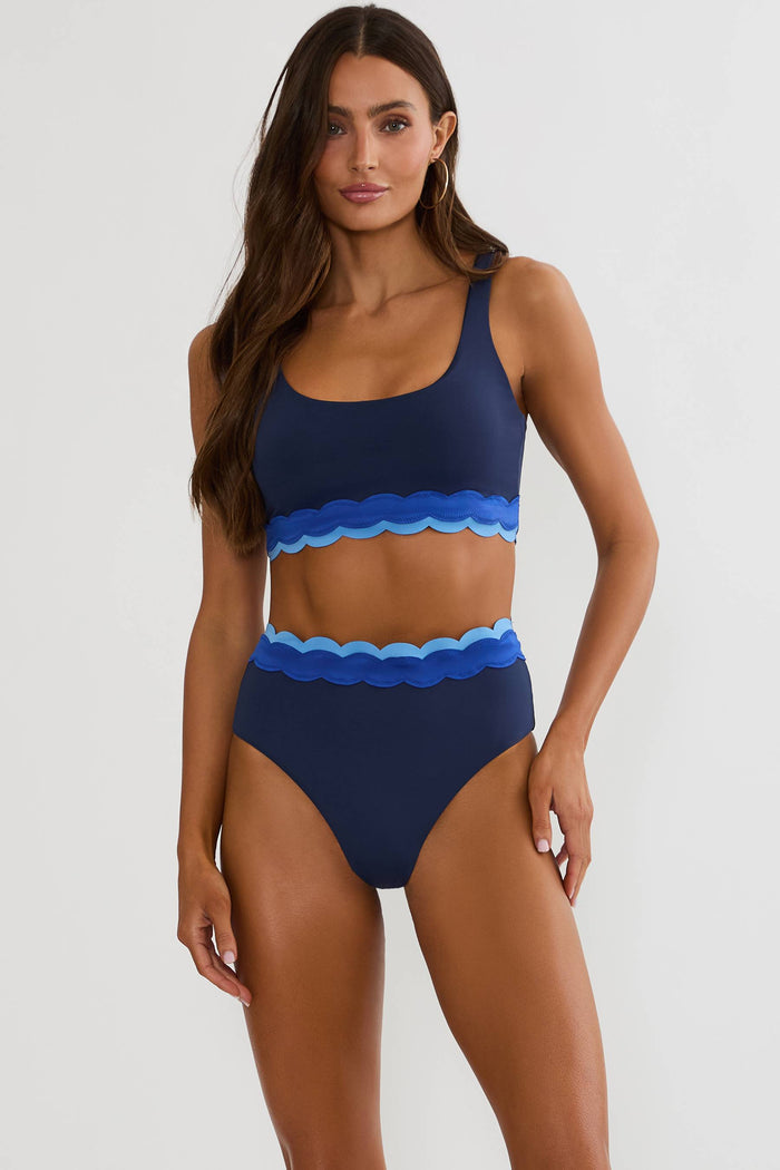 Woman wearing navy blue swimsuit with shades of blue scalloping, on a white wall.
