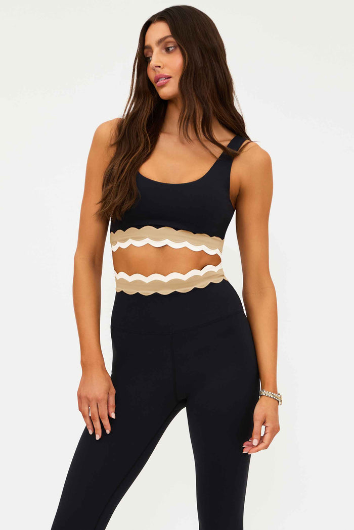 Woman wearing a black sleeveless top with beige scalloped hem and black pants on a white background