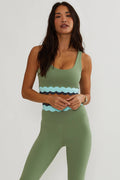 A woman modeling a sage green activewear set consisting of a scoop-neck tank top with a wavy patterned waistband and matching high-waisted leggings, against a plain light gray background.