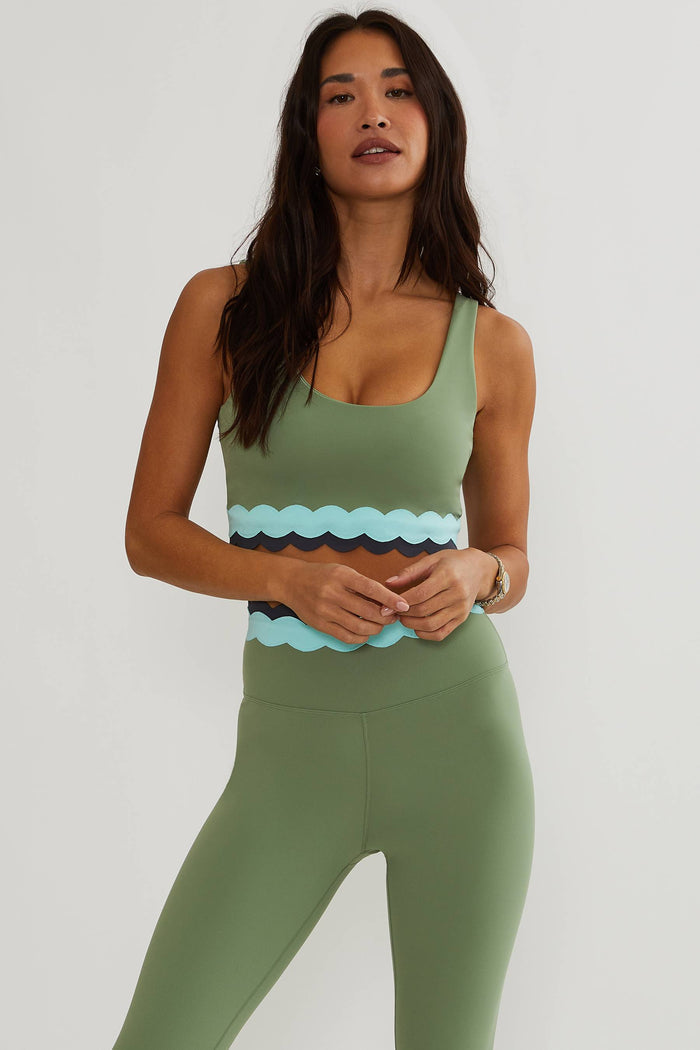 A woman modeling a sage green activewear set consisting of a scoop-neck tank top with a wavy patterned waistband and matching high-waisted leggings, against a plain light gray background.