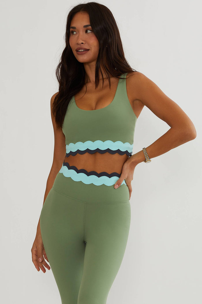 A woman modeling a matching sage green activewear set featuring a scoop-neck sports bra and high-waisted leggings with scalloped light blue and dark navy waistband details.