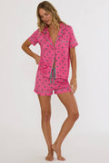 A woman modeling a two-piece pink pajama set with a small blue and white pattern, featuring a short-sleeve button-up shirt and matching shorts, against a plain white studio background.