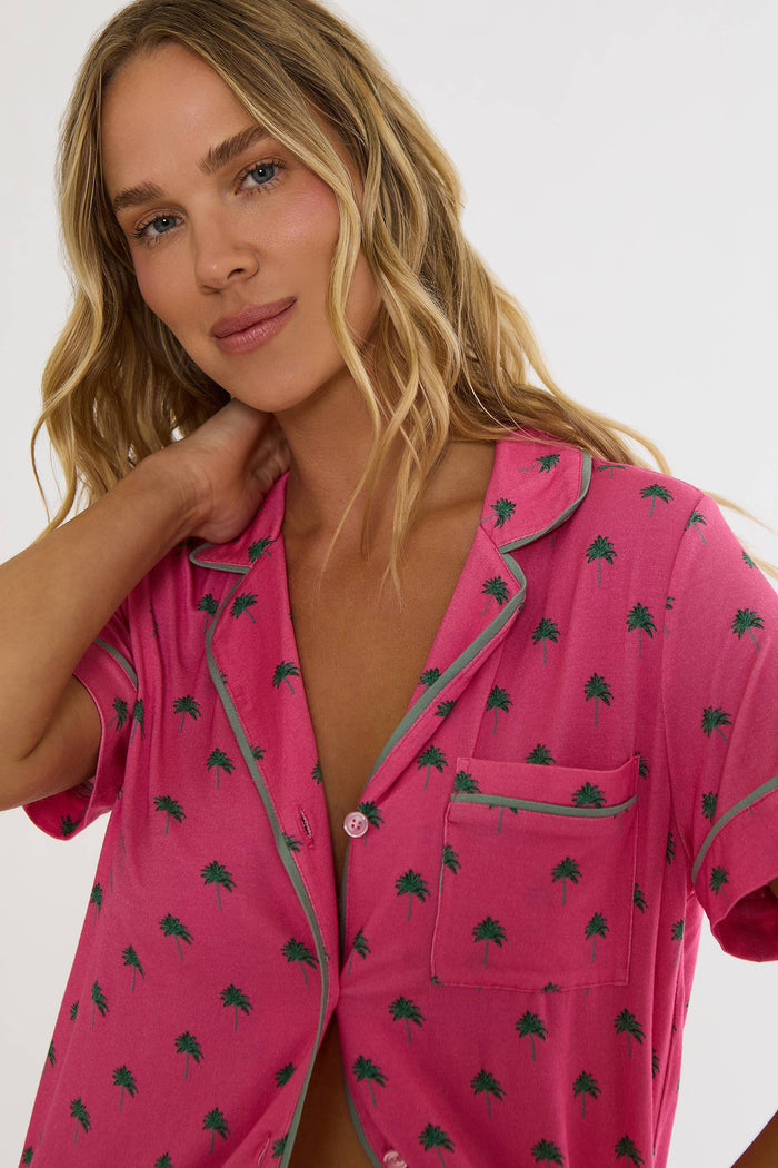 A woman wearing a pink short-sleeve pajama shirt with a dark green palm tree pattern and gray piping, shown in a close-up studio shot against a light gray background.