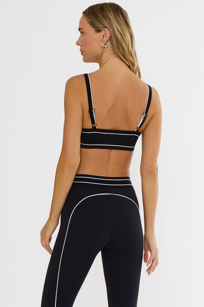 A woman shown from the back wearing a black sports bra and matching high-waisted leggings with thin white contrast piping, against a plain light gray studio background.