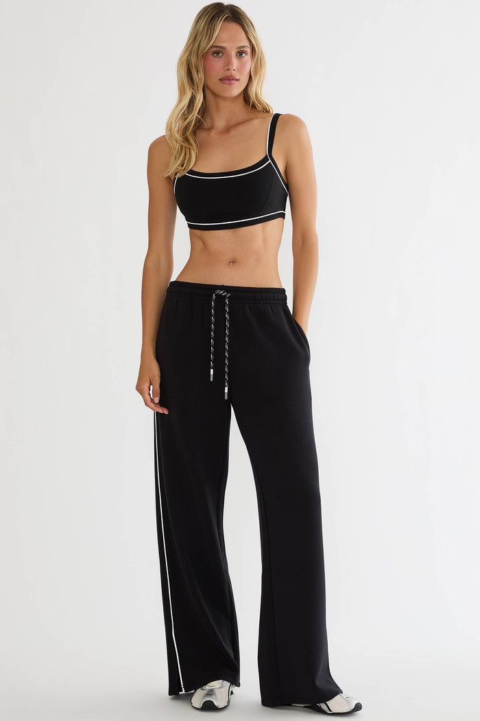 A woman modeling a black two-piece activewear set consisting of a one-shoulder crop top with white trim and matching wide-leg sweatpants with white side stripes, set against a plain light gray studio background.