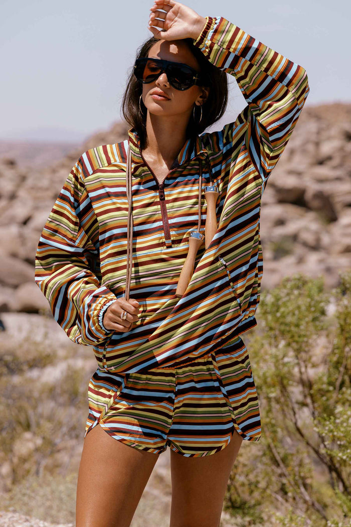 A woman wearing a matching long-sleeve quarter-zip hoodie and shorts set with a multicolor horizontal stripe pattern in brown, yellow, blue, and white, standing in a rocky desert landscape.