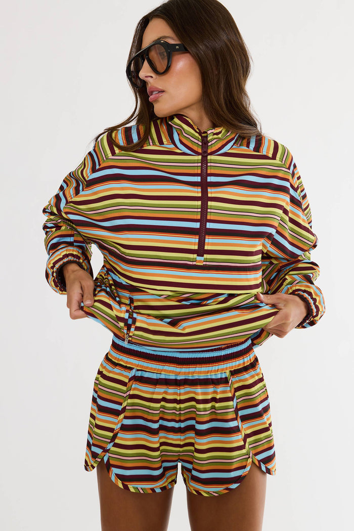 A woman modeling a matching two-piece set consisting of a long-sleeve quarter-zip hoodie and coordinating shorts, both featuring a vibrant multicolor horizontal stripe pattern in yellow, blue, brown, and white.