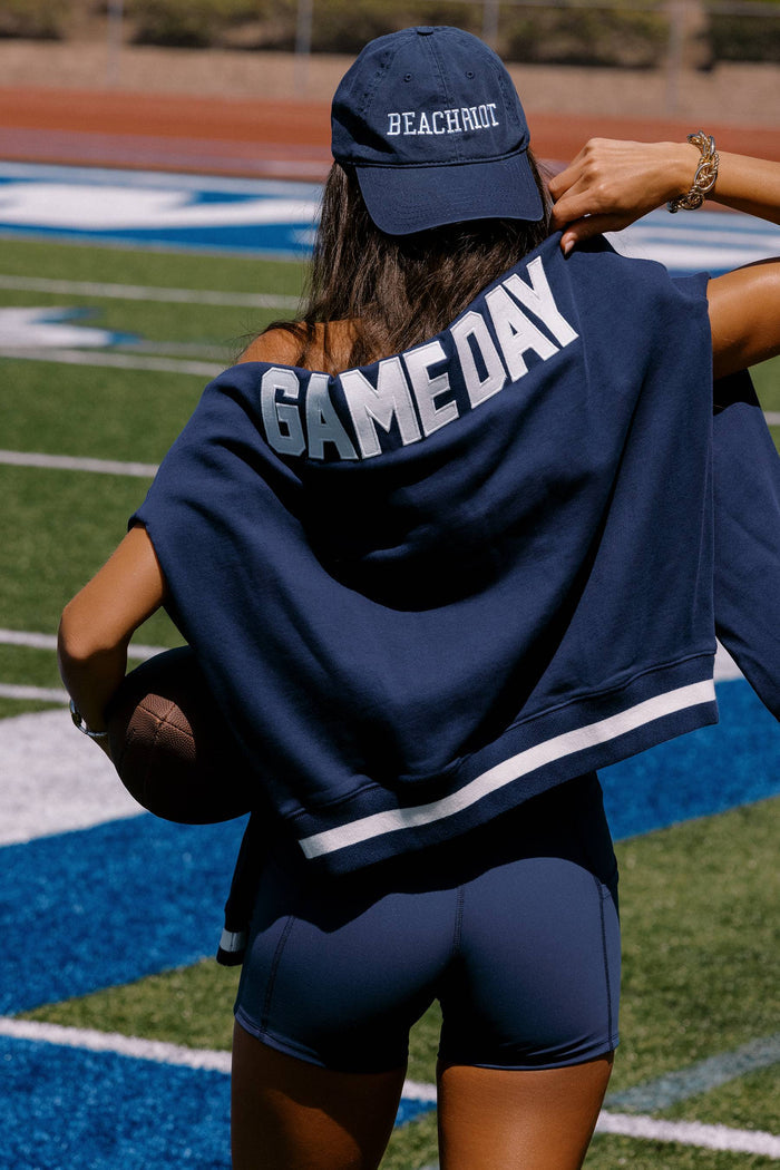 Person wearing a navy blue 'GAME DAY' jacket and cap on a sports field.